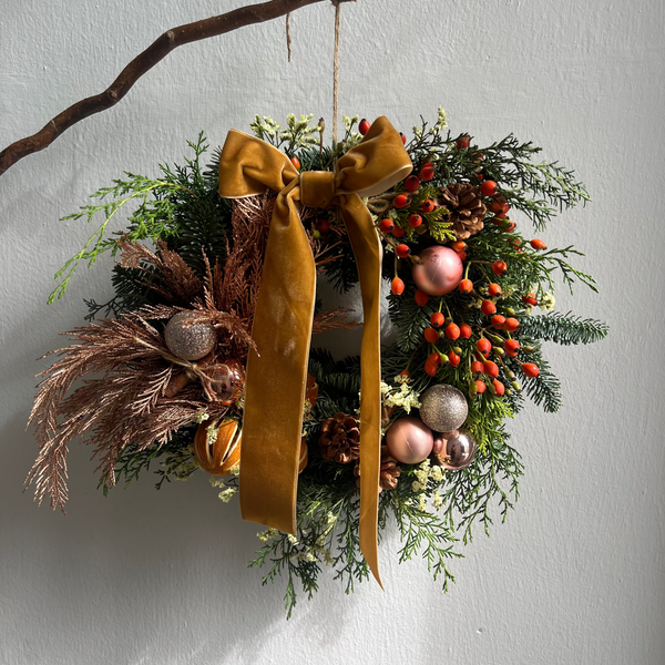 Decorative wreath with greenery, berries, and a mustard yellow ribbon on a white wall.